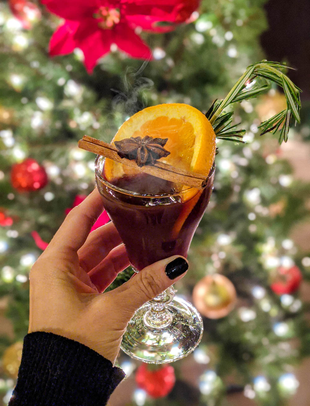 glass of hot spiced wine garnished with an orange and cinnamon stick in front of a christmas tree