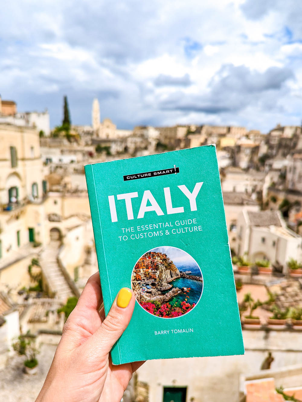 hand holding a teal Italy guidebook in front of a beige city