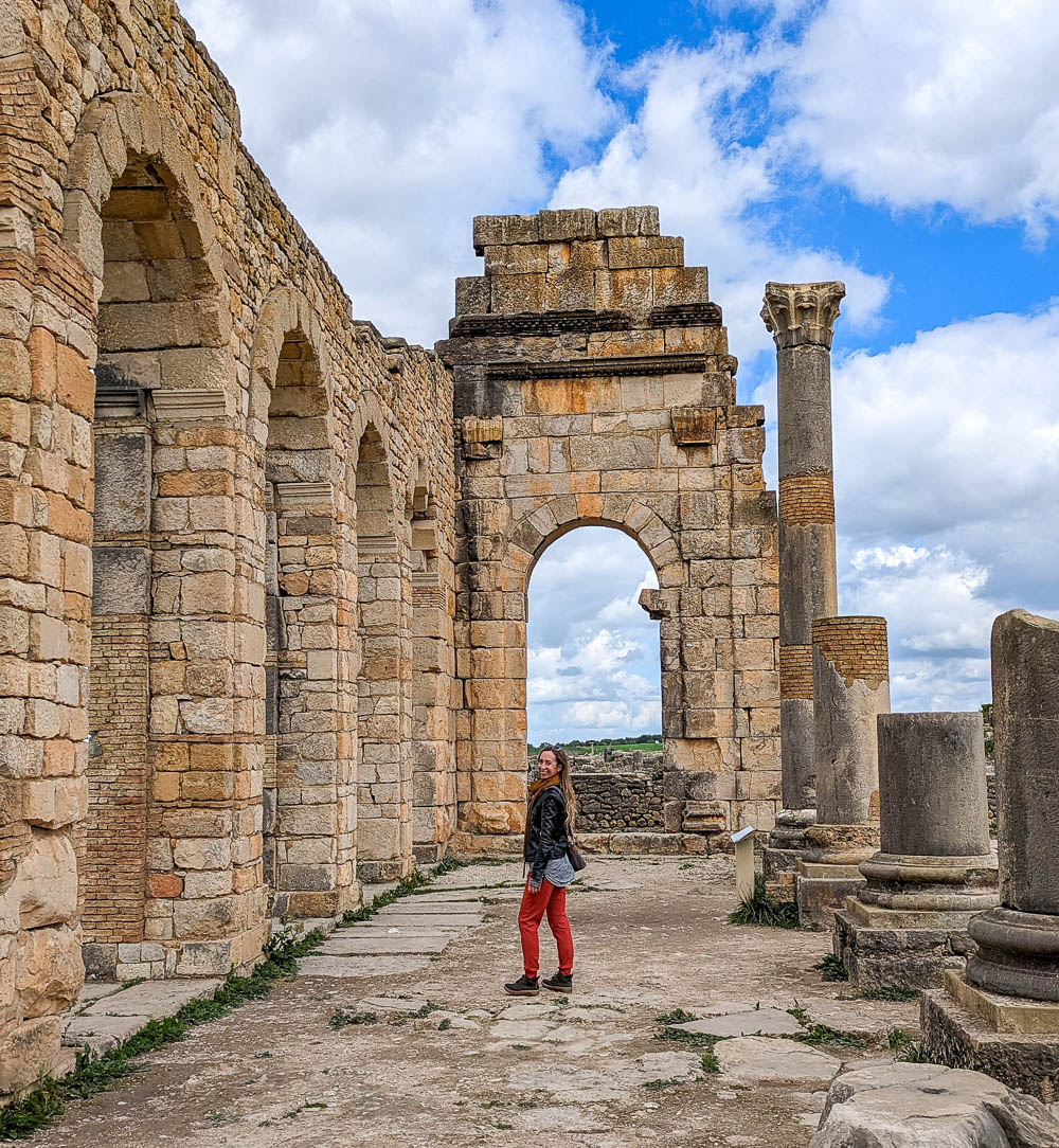 woman in red pants and black jacket standing among roman ruins