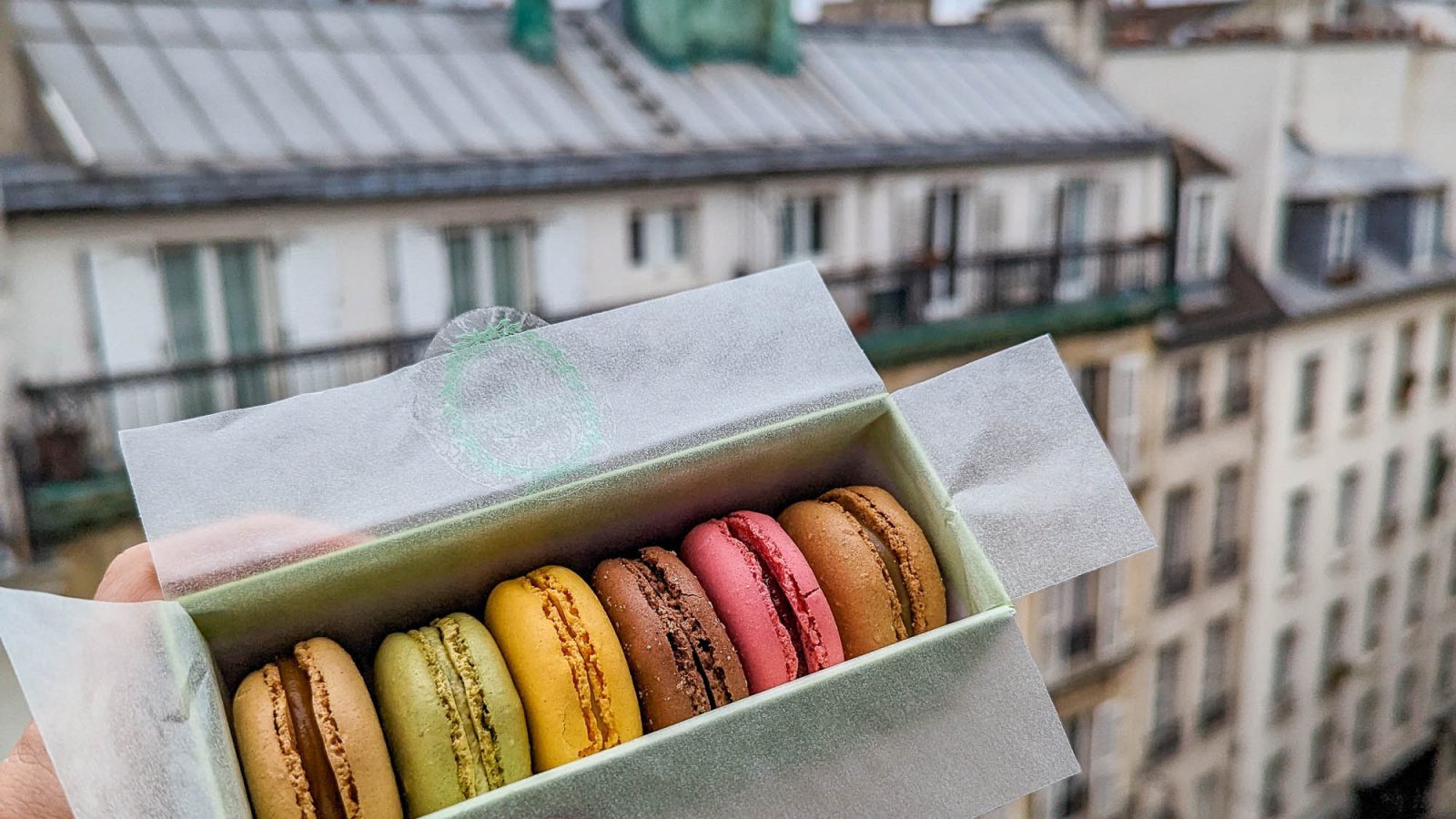 hand holding box of colorful macarons in front of a Paris skyline