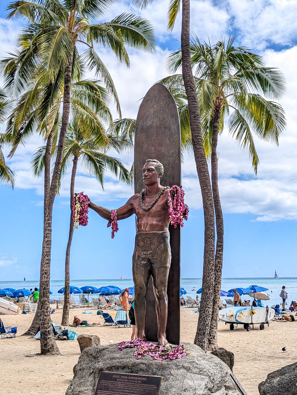 statue of a surfer that's covered in pink leis in front of a beach surrounded by palm trees