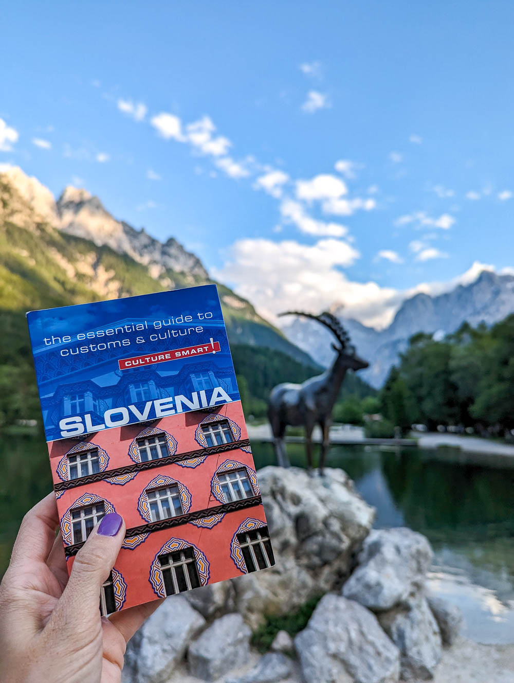 hand holding a slovenia guidebook in front of a statue of an ibex in the mountains