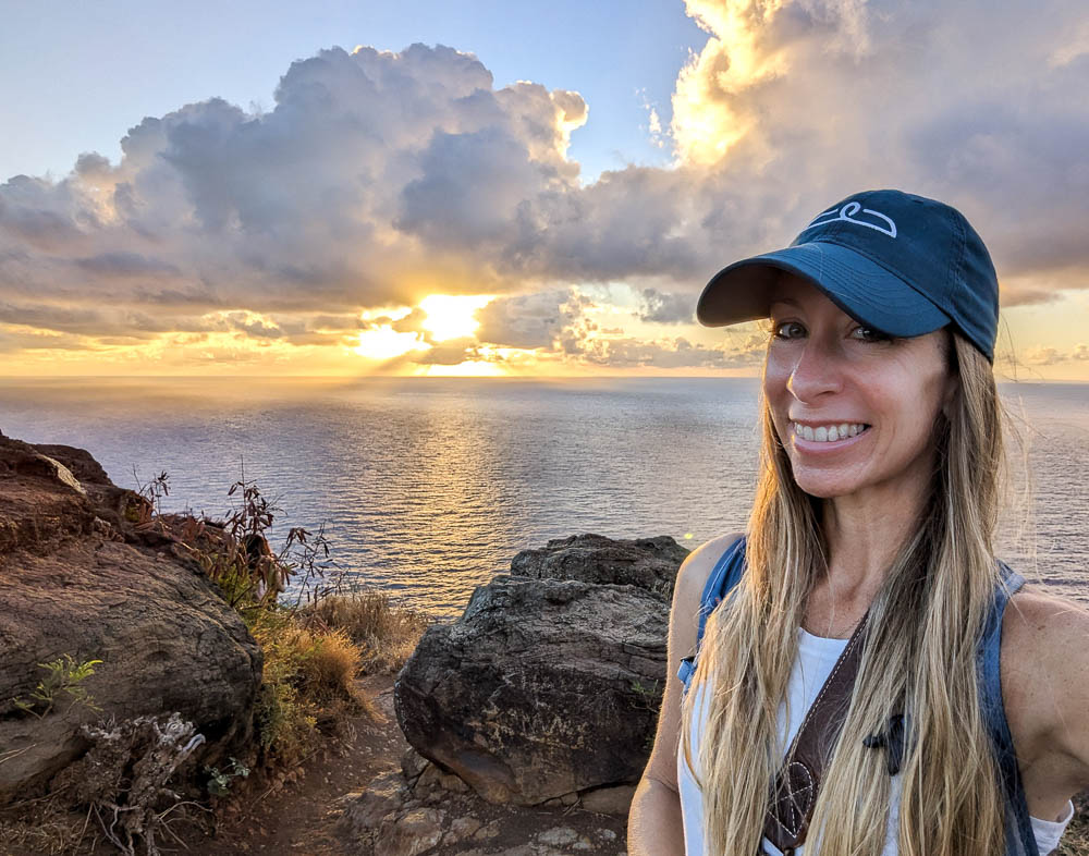 woman in dark green hat and white shirt smiling in front of a sunrise over the ocean
