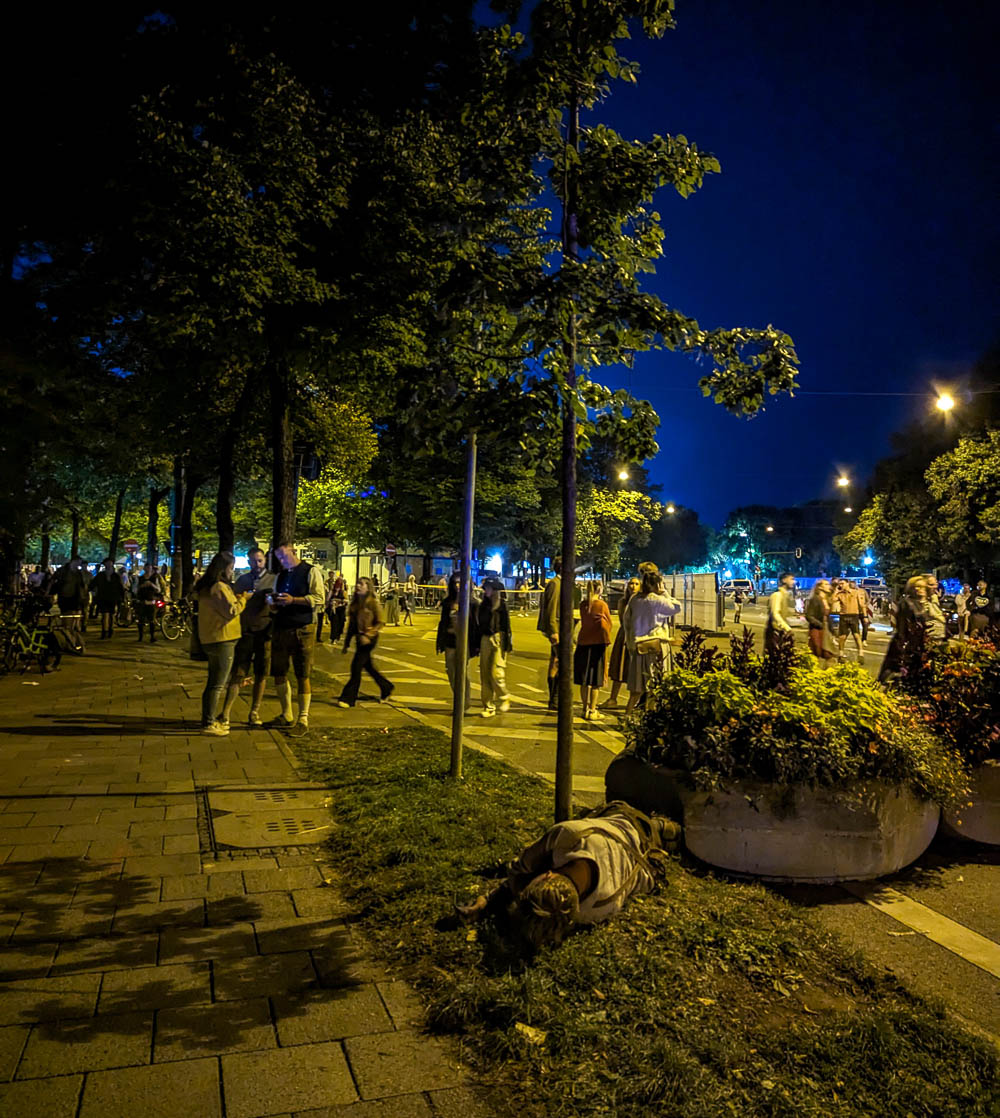 crowds of people leaving Oktoberfest at night, one man is asleep on the curb