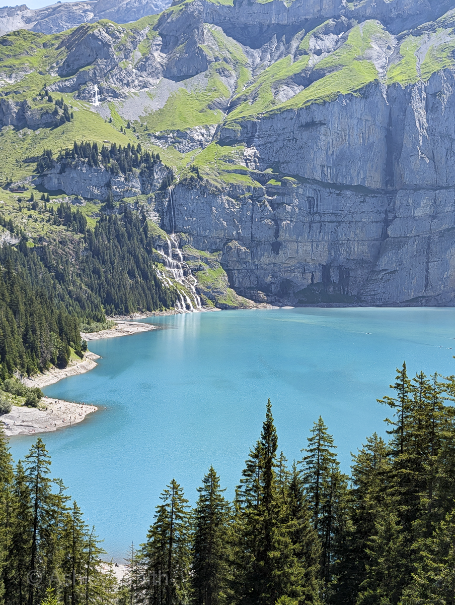 turquoise lake surrounded by green meadows and snowy mountains with waterfall